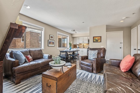 A well-lit living room with brown leather sofas, a wooden chest coffee table, and a patterned rug. The dining area and kitchen are visible in the background, with windows providing natural light.