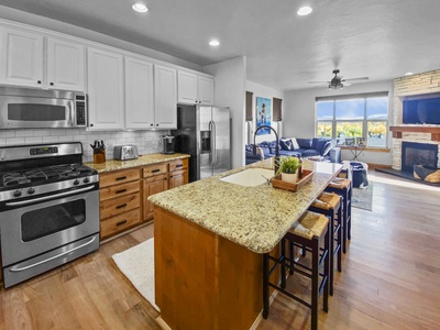 Modern kitchen with granite countertops, a central island with stools, stainless steel appliances, white cabinets, and open floor plan leading to a living area with large windows and a fireplace.