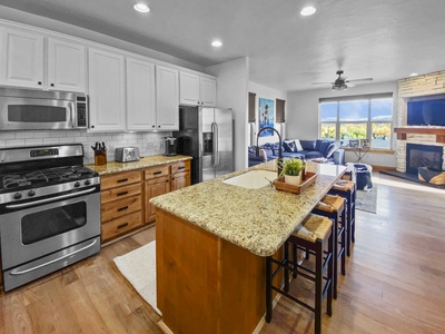 Modern kitchen with granite countertops, a central island with stools, stainless steel appliances, white cabinets, and open floor plan leading to a living area with large windows and a fireplace.