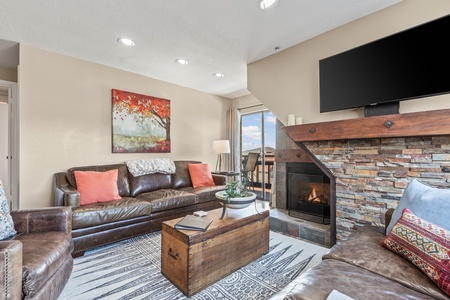 A cozy living room with a brown leather sofa set, a wooden chest coffee table, a stone fireplace, and a wall-mounted TV. A large window and artwork featuring a red tree add color to the neutral decor.