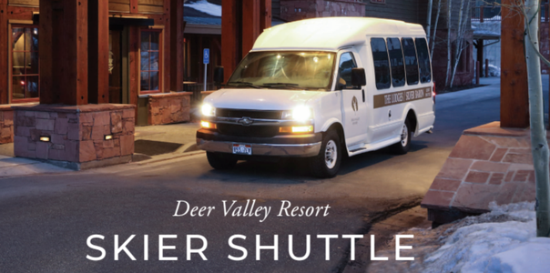 A white passenger shuttle labeled "Deer Valley Resort Skier Shuttle" is driving on a road near a stone building at dusk.