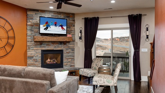 A living room with a stone fireplace, a mounted TV showing a skiing scene, brown sofa, two patterned chairs, a small table, large clock on an orange wall, and a sliding glass door with outdoor view.