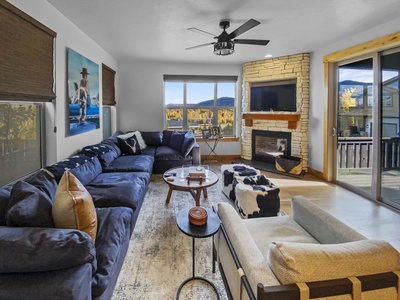 A modern living room with a large sectional sofa, armchair, coffee table, and a fireplace. Wall art, books, and decor items are arranged around the room. Large windows and sliding doors overlook a mountain view.