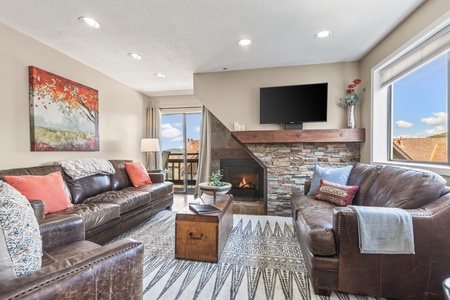 A cozy living room featuring two leather sofas, a fireplace with a stone surround, a wall-mounted TV, a wooden coffee table, and a colorful wall painting. Large windows provide natural light.