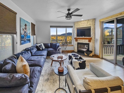 Spacious living room featuring a large sectional sofa, armchair, fireplace, mounted TV, and panoramic window views. Decor includes a landscape painting, cowhide ottomans, and a central coffee table.