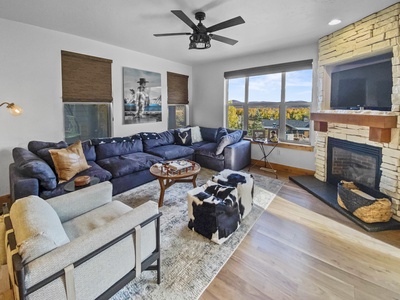 A cozy living room with a large sectional sofa, armchair, fireplace, coffee table, and large window with a scenic view. Two wall art pieces and a ceiling fan are also visible.