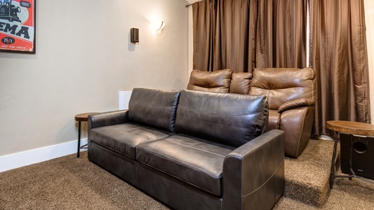 A home theater room with brown leather seating, including a couch and two recliners. The walls are beige, and a movie poster is hanging on the left side. Brown curtains cover the windows.