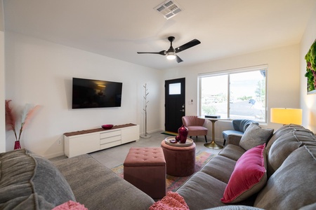 A modern living room with a gray sectional sofa, pink cushions, a wall-mounted TV, a window with daylight, a round table, and decor items including a potted plant and a coat rack.