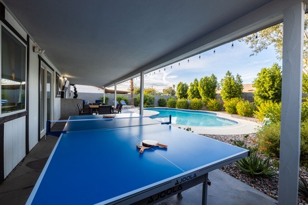 Covered patio area with a blue table tennis setup, overlooking a swimming pool and garden. Patio includes seating and string lights overhead. Background features a wall and a clear sky.