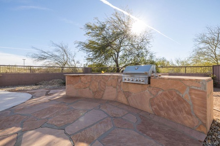 An outdoor kitchen with a built-in grill and a stone countertop sits on a flagstone patio. Trees and a fence are in the background under a sunny, clear sky.