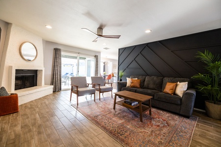 A modern living room with a dark accent wall, gray couch, wooden coffee table, two gray chairs, a rug, and a fireplace with a round mirror. Sliding glass doors lead to an outdoor patio.