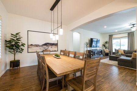 A dining area with a wooden table and chairs, modern lighting, and a large landscape painting on the wall. Adjacent living room with a TV, sectional sofa, and large windows covered with curtains.