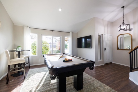 A room with a pool table in the center, a TV mounted on the wall, a small table with two chairs, a carpet on a wooden floor, and large windows letting in natural light.