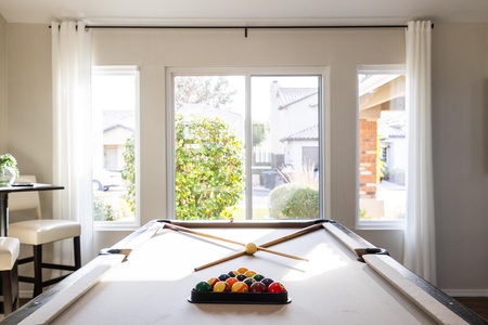 A pool table in a bright room with large windows showing an outdoor view. The table has a triangle of colorful balls and four pool cues arranged in an X pattern. Two chairs are visible on the left.