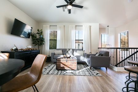 Bright living room with large windows, a wall-mounted TV, two grey armchairs, and a cushioned trunk as a coffee table. Wooden floor, patterned rug, and indoor plant enhance the modern decor.