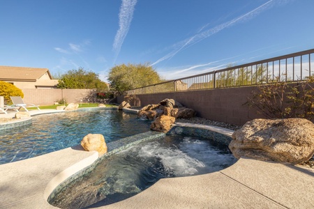 A backyard pool with an attached hot tub, surrounded by rocks and a concrete patio, is shown under a clear, blue sky with some contrails. A fence and neighboring houses are visible in the background.