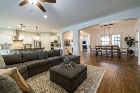 A spacious living room with a large grey sectional sofa, a patterned rug, and wooden flooring. The room opens to an adjacent dining area with a wooden table and hanging lights, and a modern kitchen area.