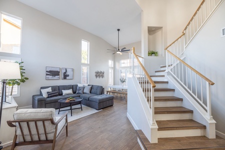 A modern living room with a gray sectional sofa, a wooden chair, and a coffee table. Stairs with white railings lead to the upper floor. The room is well-lit with natural light from large windows.