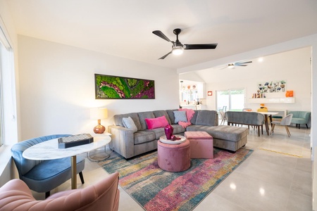 Modern living room with a gray sectional sofa, colorful rug, pink ottoman, round marble table, and ceiling fan. Art decorates the walls and there is a view into an adjoining dining area.