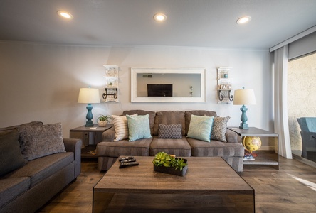 A modern living room with a gray sofa, a coffee table with a plant, two lamps with blue bases, a wall mirror, and decorative pillows. A second sofa is partly visible on the left.