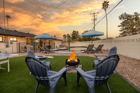 A backyard features a fire pit with four chairs, a swimming pool with lounge chairs and umbrellas, and a house in the background, all set under a vibrant sunset sky.