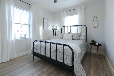 A modern bedroom with a black metal bed frame, patterned bedspread, two windows with white curtains, a small bedside table with a clock, and a dresser with a lamp and framed picture.