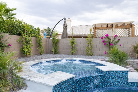 A backyard with a small, round pool featuring a stone deck and blue mosaic tiles. There are flowering plants and a lounge chair in the background. The area is fenced with a brick wall.