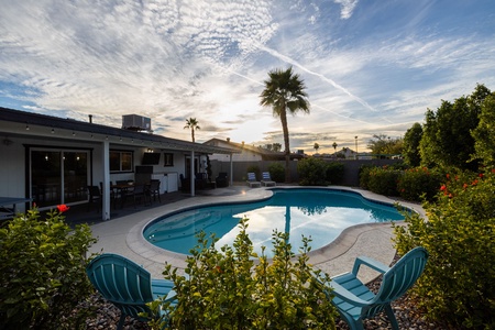 A backyard with a round swimming pool, patio area, blue chairs, lush greenery, and a palm tree under a partly cloudy sky at sunset.