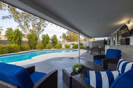 A covered patio with wicker furniture and blue cushions overlooks a backyard pool surrounded by greenery. A dining table and a TV are also visible on the patio.