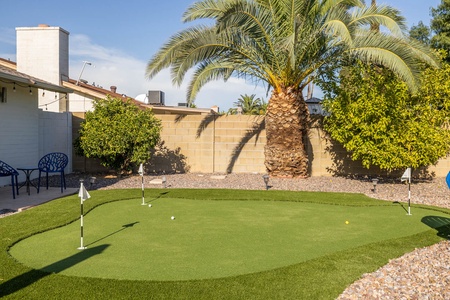 A backyard putting green with four holes, surrounded by gravel and trees, including a large palm tree. Two blue chairs are placed on a concrete patio area in the background.