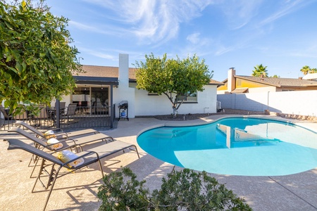 A backyard with a swimming pool, four lounge chairs, a tree, and a covered patio area with outdoor furniture near a single-story house under a clear blue sky.