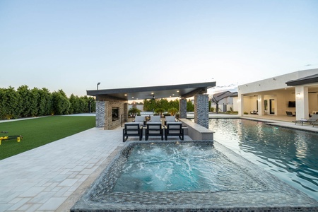 An outdoor space features a swimming pool, attached hot tub, covered patio area with a dining table, and a large green lawn surrounded by tall hedges. Homes are visible in the background.