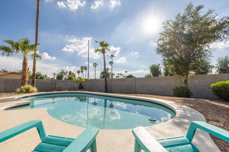 A clear swimming pool with turquoise chairs on the deck, surrounded by palm trees and a stone wall, under a sunny sky.