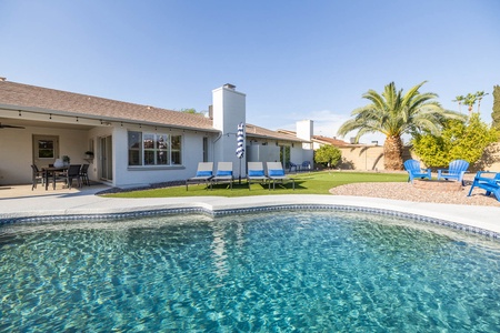 A backyard with a swimming pool, patio area with a table and chairs, lawn chairs near a small fireplace, and palm trees in the background.