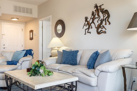 A living room with white leather sofas, blue cushions, and a marble coffee table. The room features wall art of dancing figures and a round mirror, with a potted plant on the coffee table.