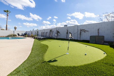 Outdoor putting green with several golf balls and flags, next to a concrete path, lawn chairs, and a swimming pool under a clear blue sky.