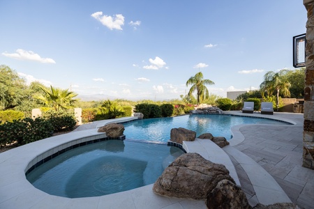 A swimming pool with a spa area, surrounded by rocks and greenery, next to a patio with two white lounge chairs. The scene is set under a clear, blue sky with some clouds and distant mountains visible.