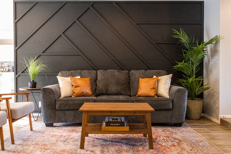 A gray sofa with white and orange pillows is set against a black geometric accent wall. A wooden coffee table sits on a patterned rug, with potted plants on each side of the sofa.