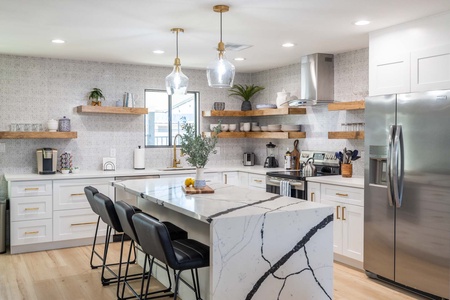 Modern kitchen with white cabinets, marble island, black bar stools, stainless steel appliances, open wooden shelves with dishes, and pendant lights.