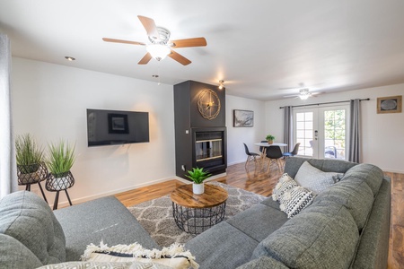 Modern living room with a gray sectional sofa, round wooden coffee table, wall-mounted TV, and fireplace. Dining area with table and chairs is visible through an arched entryway. Ceiling fans on.