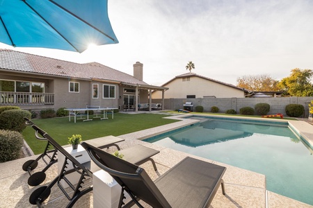 A backyard with a pool, sun loungers, a table tennis setup, and a house with a patio. There's an umbrella providing shade, and another house and a tree are visible over the fence.