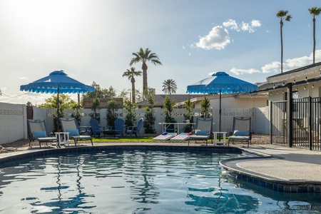 A backyard pool area with blue umbrellas, lounge chairs, and a grassy area with a table and chairs. Tall palm trees and a white fence are in the background. The sun is shining brightly.