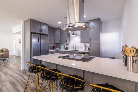 Modern kitchen with gray cabinets, stainless steel appliances, a central island with a stovetop, and three black barstools. The floor is wood, and the walls are white. Living area visible in the background.