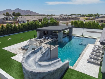 A modern backyard with a swimming pool featuring a waterslide, a covered lounge area, and surrounding green artificial turf. There are lounge chairs by the pool and houses and mountains in the background.