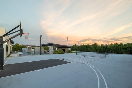 Outdoor basketball court with two hoops, surrounded by greenery, under a sky with warm sunset hues. A tall structure with roof is visible in the background.