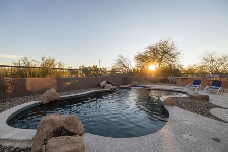 A serene outdoor swimming pool with a few large rocks around the edge, surrounded by a stone patio with two lounge chairs, and trees in the background under a clear sky with the sun setting.