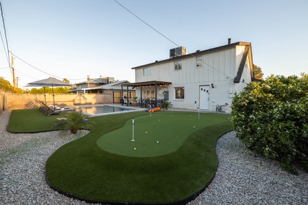 A backyard with a putting green beside a swimming pool, outdoor seating under a pergola, and a two-story house in the background.