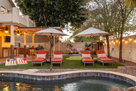 A backyard features a pool in the foreground, four orange lounge chairs with umbrellas, a cornhole game, outdoor dining area, and a small putting green, all surrounded by trees and string lights.