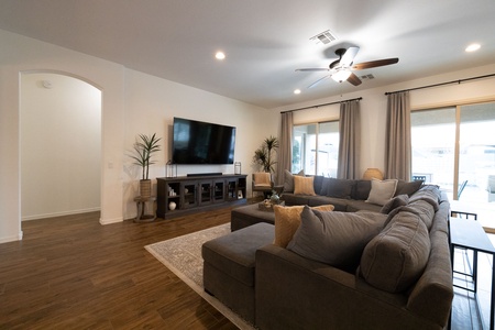 A living room with a large gray sectional sofa, a TV mounted on the wall, ceiling fan, hardwood floors, and sliding glass doors leading outside. The room features potted plants and a small area rug.