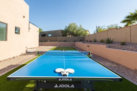 A blue JOOLA ping pong table with paddles and balls on it is set up in an outdoor area with a beige wall, gravel, plants, and clear sky in the background.